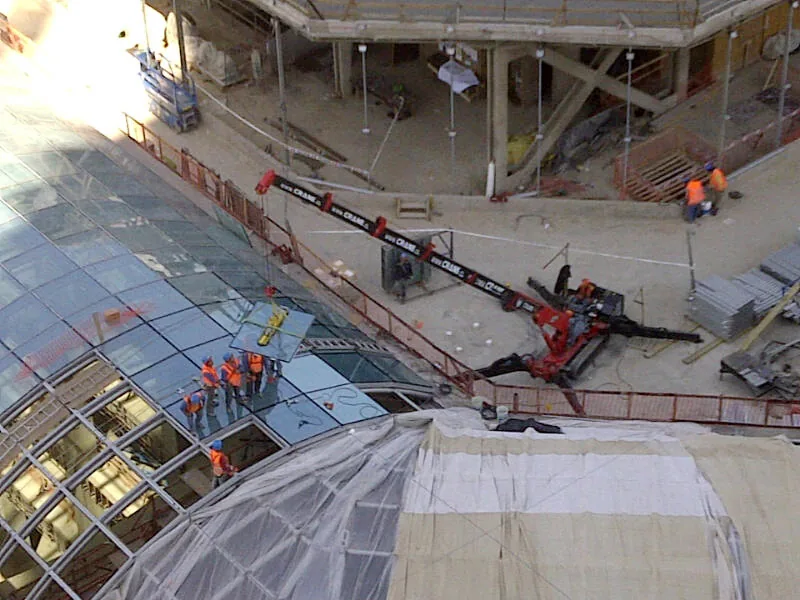 Vista desde lo alto: la minigrúa araña trabajando al borde de una cúpula de vidrio, con operarios repartidos sobre la estructura que dan la escala del vano