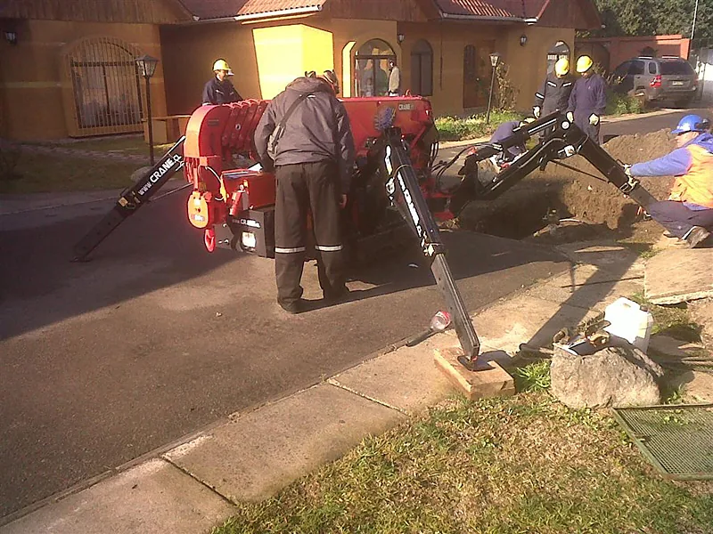 La minigrúa araña operando al atardecer en el acceso de una vivienda, con los estabilizadores apoyados sobre el pavimento y dos operarios guiando la carga