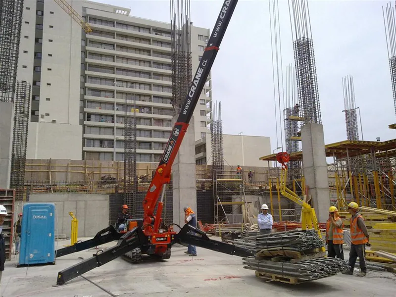 La minigrúa araña izando en una obra rodeada por la cuadrilla, con enfierradura apilada en el suelo y un edificio de varios pisos en construcción al fondo
