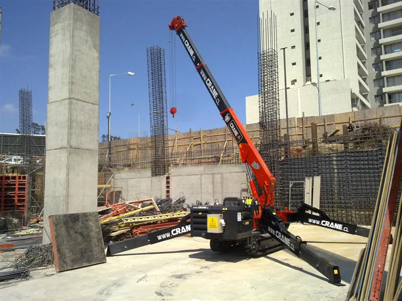 La minigrúa araña con sus cuatro estabilizadores desplegados sobre la losa de una obra, la pluma extendida en diagonal contra el cielo despejado y una columna de hormigón recién descimbrada en primer plano