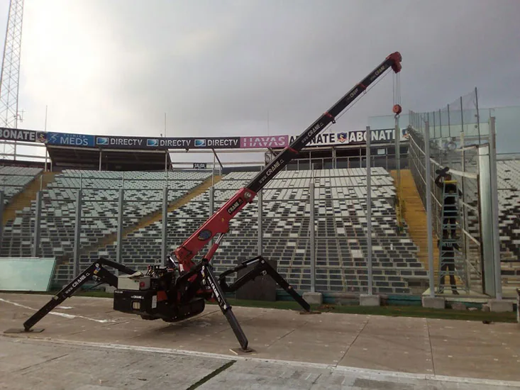 La minigrúa araña con las cuatro patas desplegadas sobre la pista de un estadio, frente a las graderías vacías