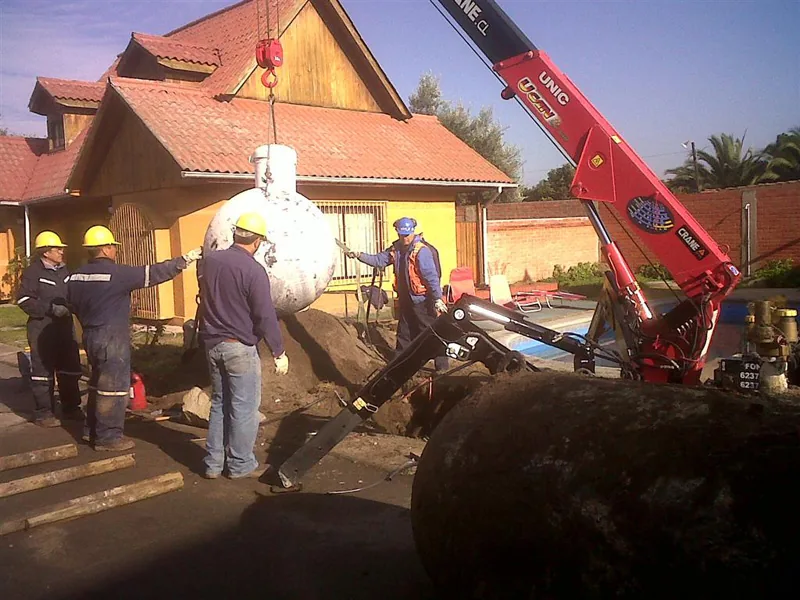 Dos operarios guiando un estanque cilíndrico que la minigrúa araña baja hacia una excavación, en el patio de una vivienda