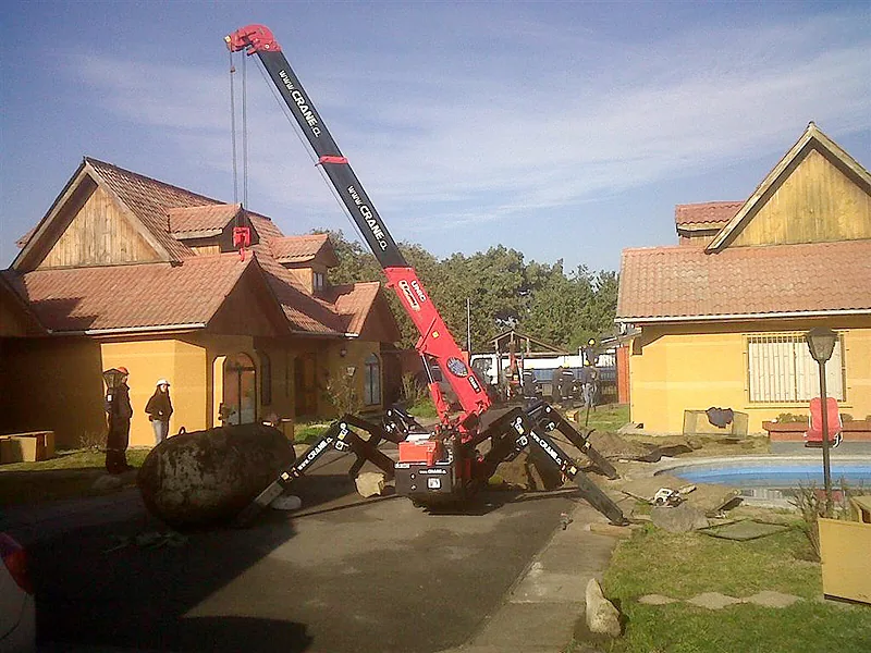La minigrúa araña con la pluma extendida sobre el techo de una vivienda, rotulada www.crane.cl en el brazo, trabajando entre las casas de un condominio