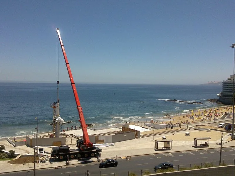 La grúa de 100 toneladas trabajando en el borde costero, con la pluma extendida sobre el paseo y la playa llena al fondo
