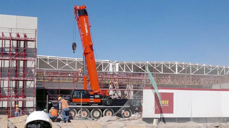 La grúa de 100 toneladas montando una estructura metálica curva de techumbre, con la cuadrilla trabajando abajo