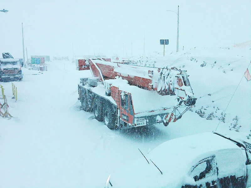 La grúa telescópica de 55 toneladas cubierta de nieve en una faena de cordillera, con la pluma recogida y la ventisca borrando el horizonte