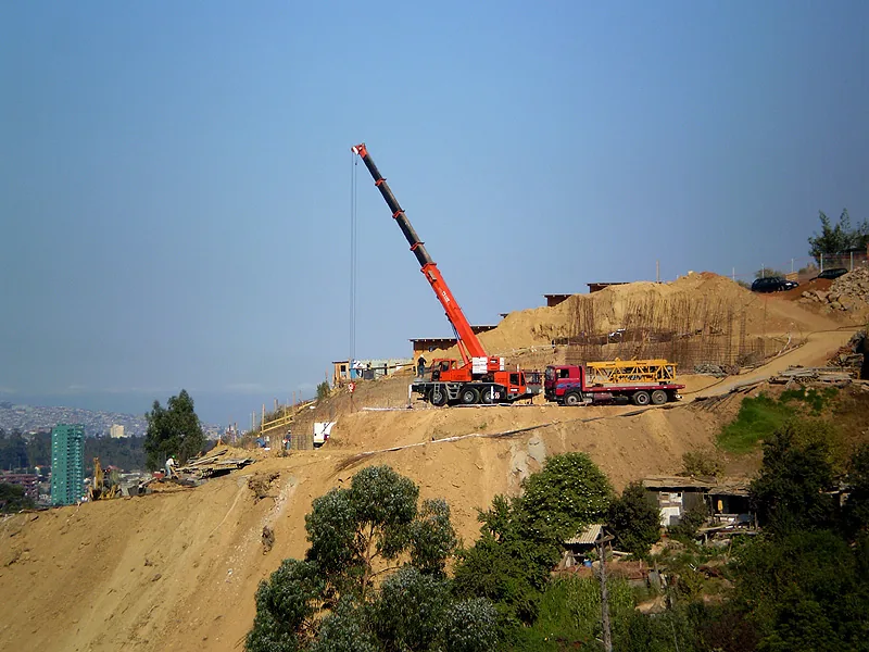 La grúa de 55 toneladas izando sobre una plataforma de tierra excavada en ladera, con camiones de apoyo al lado y la ciudad al fondo