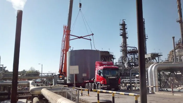 La grúa de 80 toneladas colocando un estanque cilíndrico en una planta industrial, con las chimeneas y el trazado de cañerías detrás