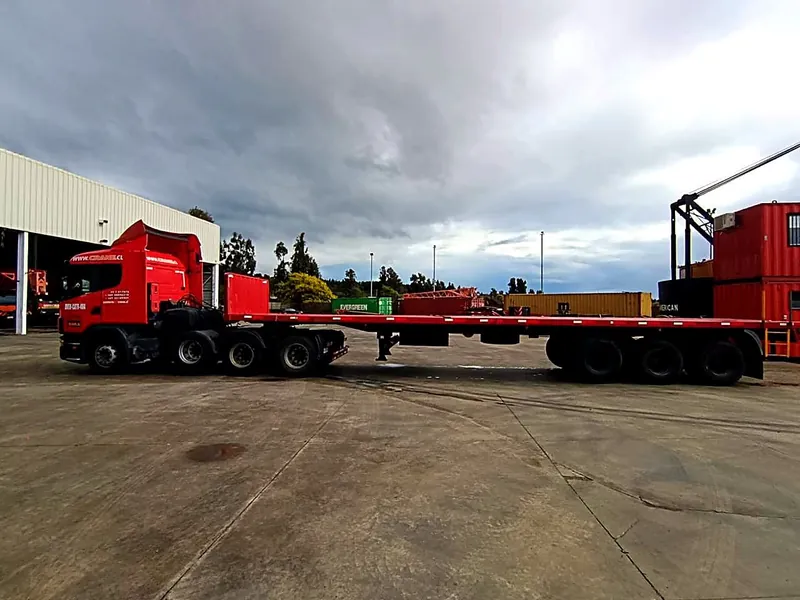 La rampla plana de 22 toneladas vista de perfil, con la plataforma vacía en todo su largo y los tres ejes atrás, enganchada al tractocamión en un patio con contenedores